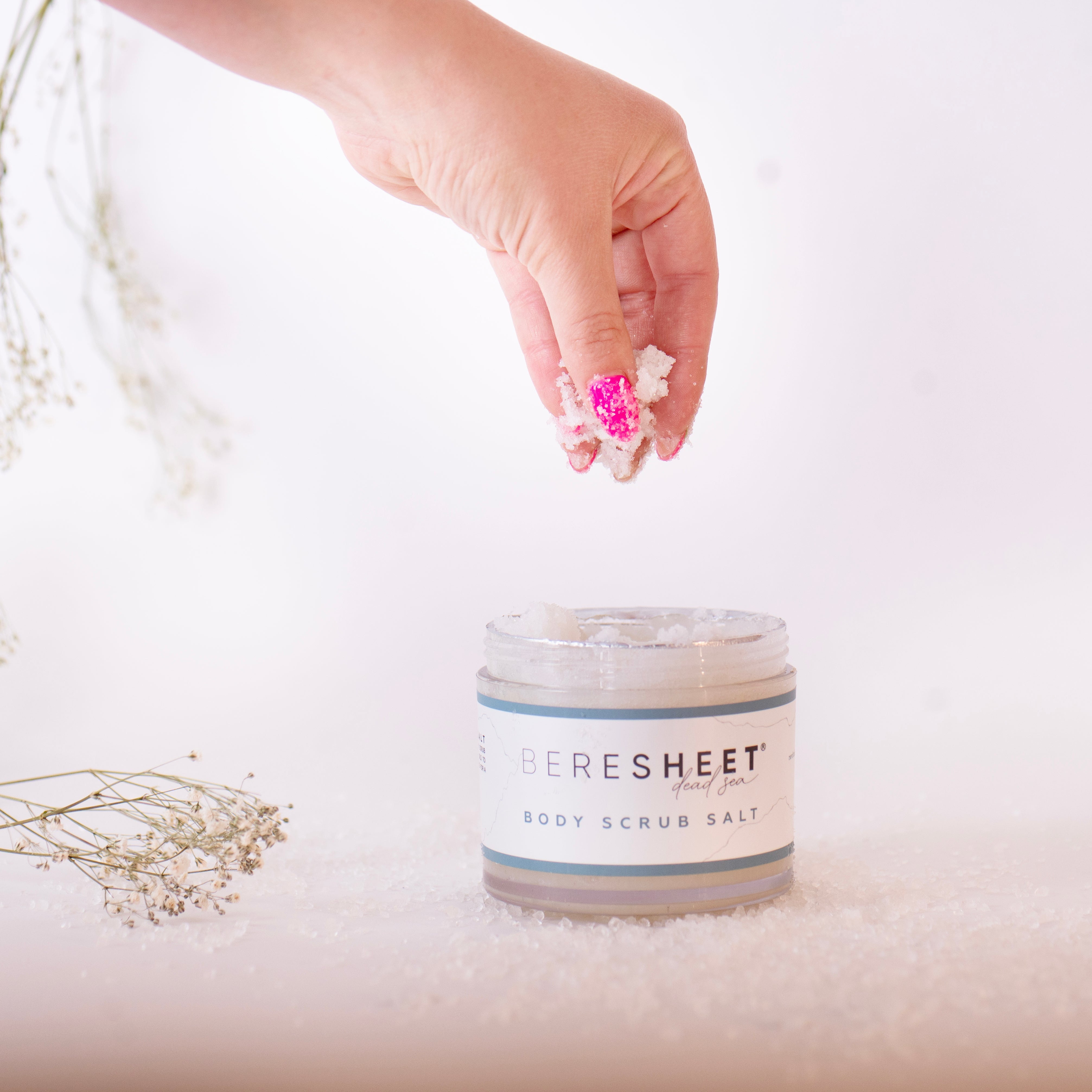 Hand holding flowers above a jar of 'PERESHEET' body scrub salt on a light background