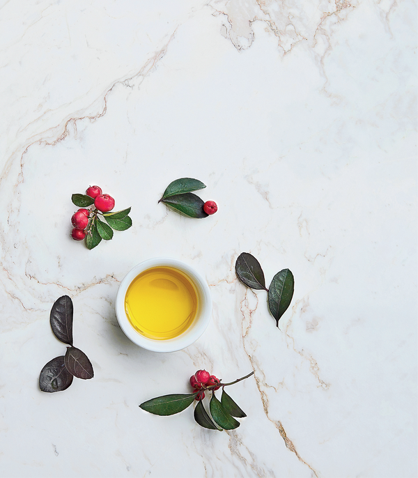 Yellow liquid in a white bowl surrounded by red berries and green leaves on a marble surface