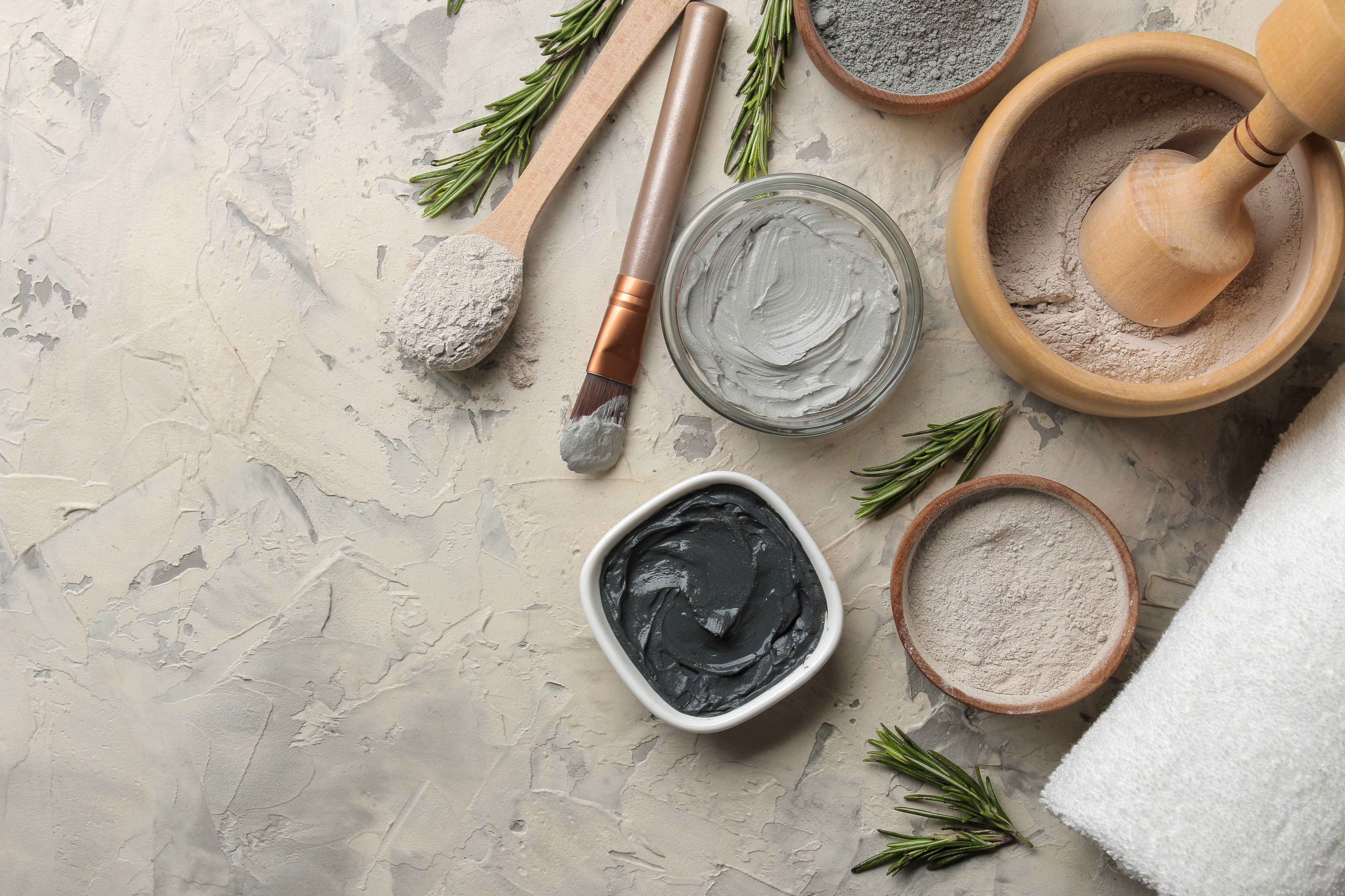 Cosmetic ingredients including a bowl of gray clay, black charcoal, and white powder on a light stone surface.